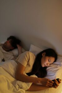 A couple lies in bed, emotionally distant, with one person scrolling on their phone. Seeking support from a Fort Worth trauma therapist can help rebuild connection. Trauma therapy in Fort Worth, TX offers guidance in healing intimacy struggles caused by past wounds.